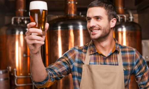 Happy Brewer. Happy young male brewer in apron holding glass with beer and looking at it with smile while standing in front of metal containers
